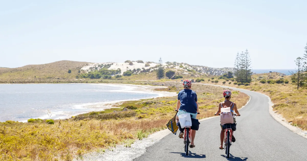 Two cyclists riding a quiet road past a pale salt lake and low dunes on Rottnest Island