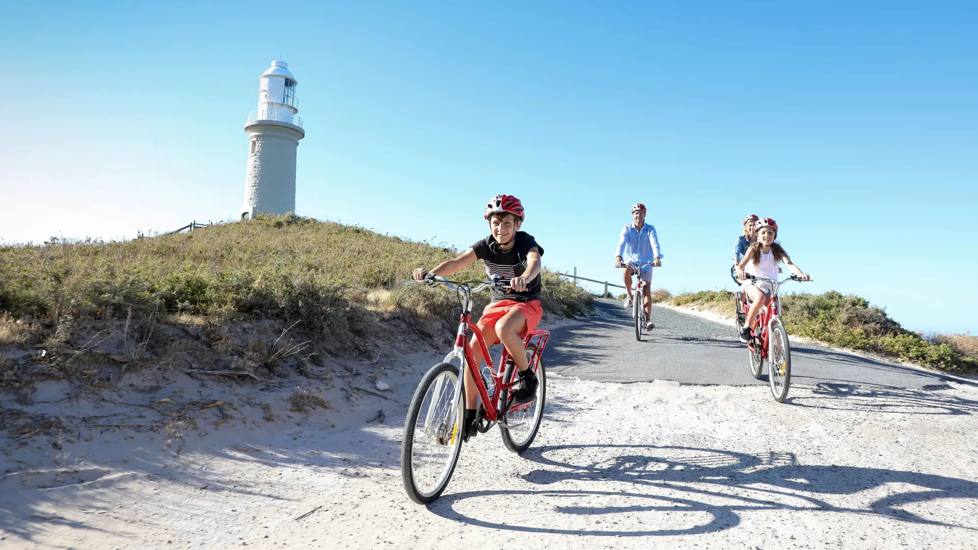 Cyclists riding a sandy road past a white lighthouse on a dune on Rottnest Island, from the ferry and bike day tour listing